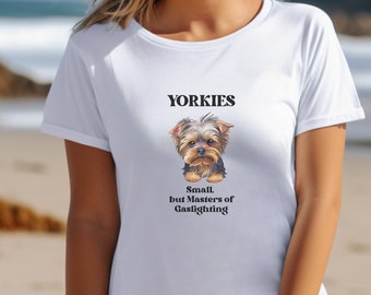 Op de afbeelding: Wit T-shirt met het woord "Yorkies" en een illustratie van een Yorkshire Terrier. De tekst "Small, but Masters of Gaslighting" staat eronder. De persoon draagt het T-shirt op een strand met de zee op de achtergrond.