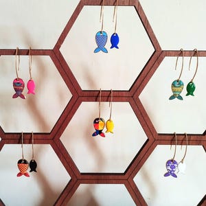 May include: A display of colourful fish-shaped earrings on a hexagonal wooden stand. The earrings are made of enamel and have gold hooks. The fish are painted in various patterns and colours, including blue, green, pink, yellow, and black.