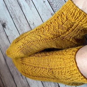 Könnte beinhalten: Ein Paar senfgelbe, handgestrickte Socken mit einem strukturierten Zopfmuster, das vertikal über die gesamte Länge verläuft. Die Socken verfügen über ein geripptes Bündchen am oberen Rand und ein gleichmäßiges Strickmuster durchgehend.