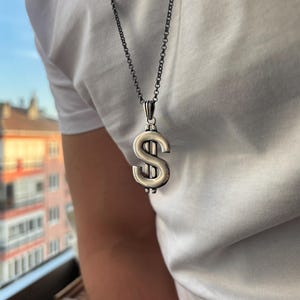 Op de afbeelding: Een zilverkleurige dollarteken-hanger hangend aan een donkere, dunne metalen ketting. De hanger heeft een gepolijste, gladde afwerking met een verticale lijn door het midden van het valutasymbool, gedragen op een effen wit t-shirt.