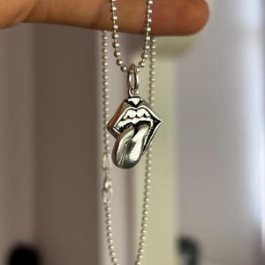Pode incluir: Um pingente de metal prateado em forma de lábios com uma língua saliente, suspenso por um colar de corrente de bolinhas a condizer. O pingente apresenta detalhes pretos em baixo-relevo para definir os contornos dos lábios e da língua.