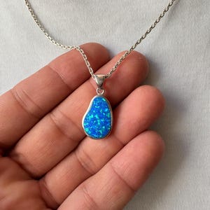 Puede incluir: Un collar de plata con un colgante en forma de lágrima que presenta una incrustación iridiscente de color azul vibrante. El colgante tiene un borde metálico liso y cuelga de una delicada cadena de plata, mostrando una superficie brillante.