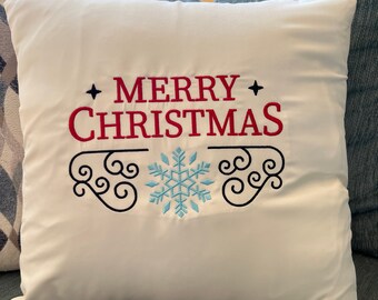 May include: Cream-colored square pillow with the embroidered text "MERRY CHRISTMAS" in red. A light blue snowflake is embroidered below the text, with black decorative flourishes. The pillow is on a blue couch.