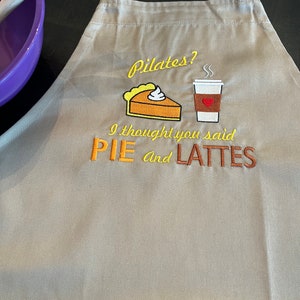 May include: A tan apron with the embroidered text "Pilates? I thought you said PIE and LATTES." The design includes a slice of pie with whipped cream and a coffee cup. The apron has adjustable straps and is made of a durable fabric.