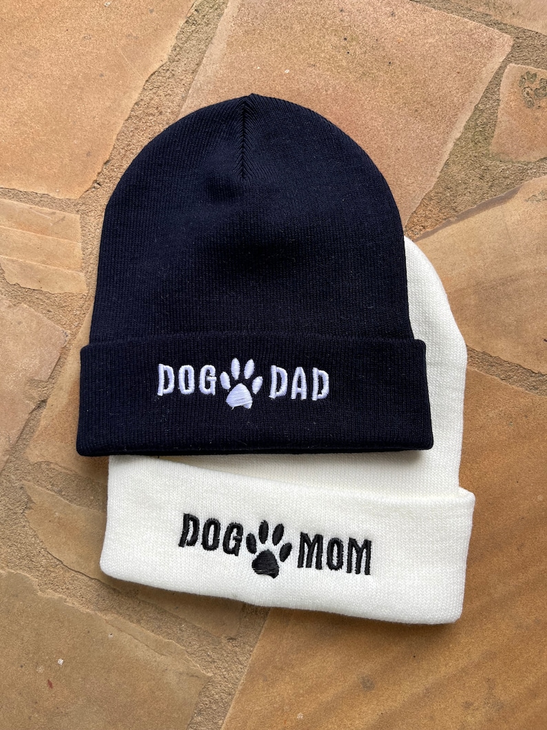 Op de afbeelding: Twee gebreide mutsen, &eacute;&eacute;n marineblauw en &eacute;&eacute;n wit. De marineblauwe muts heeft de witte borduursel "DOG DAD" met een pootafdruk. De witte muts heeft de zwarte borduursel "DOG MOM" met een pootafdruk. Beide mutsen zijn aan de rand gevouwen.