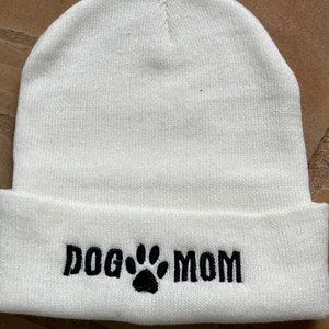 Op de afbeelding: Een witte gebreide muts met een omgeslagen rand. De tekst "DOG MOM" is in zwart geborduurd, met een pootafdruk tussen de woorden. De muts is gemaakt van een zacht, gebreid materiaal.