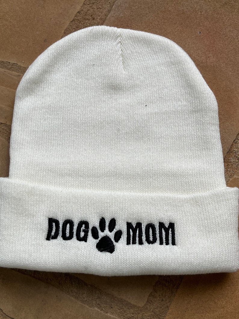 Op de afbeelding: Een witte gebreide muts met een omgeslagen rand. De tekst "DOG MOM" is in zwart geborduurd, met een pootafdruk tussen de woorden. De muts is gemaakt van een zacht, gebreid materiaal.