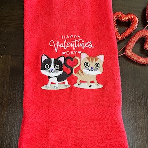 May include: Red Valentine's Day towel with embroidered cartoon cats. The towel features the text "Happy Valentine's Day" in white script above the cat design. The cats have heart-shaped tails.