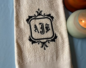 May include: Cream-colored bath towel with a black embroidered monogram within a decorative frame. The towel has a textured surface and a patterned border. Two lit candles are in the background.