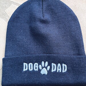 Op de afbeelding: Marineblau gebreide muts met de geborduurde tekst "DOG DAD" en een pootafdruk. Een leuk cadeau voor hondenliefhebbers.