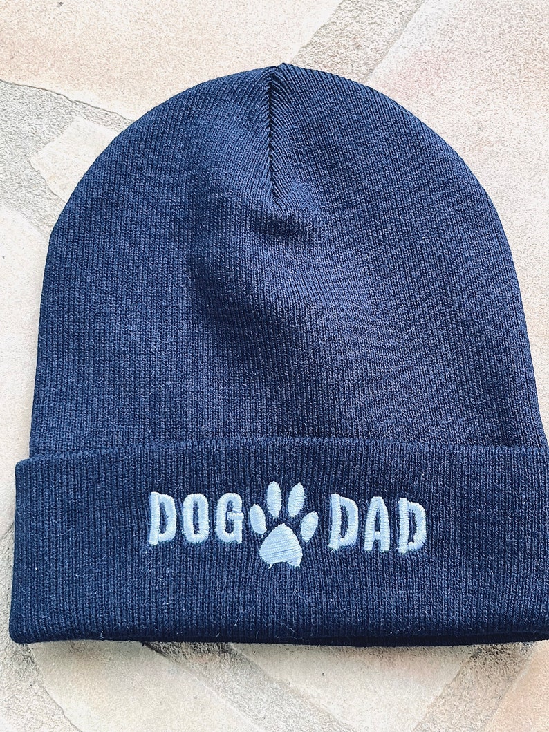 Op de afbeelding: Marineblau gebreide muts met de geborduurde tekst "DOG DAD" en een pootafdruk. Een leuk cadeau voor hondenliefhebbers.