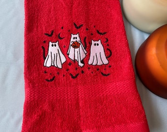 May include: A folded red hand towel with an embroidered Halloween design. The design features three cartoon ghosts, bats, and stars. One ghost is holding a pumpkin. The towel is on a light blue surface.