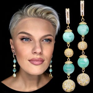 May include: Long, dangling earrings featuring alternating turquoise-colored orbs and gold-colored, crystal-studded spheres. The earrings have a gold-colored, linear top with crystal accents and are set against a black background.