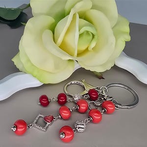 Puede incluir: Un llavero con cuentas rojas y detalles plateados. El llavero presenta múltiples hilos de cuentas rojas, con un dije en forma de corazón y un dije en forma de diamante. Una gran rosa amarilla está en el fondo.