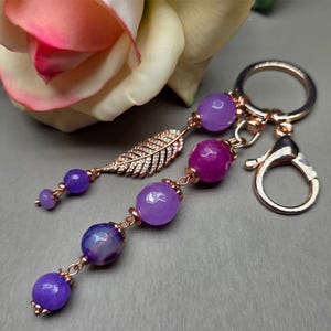 Puede incluir: Un llavero de oro rosa con un mosquetón y un anillo. El llavero presenta un dije con cuentas moradas y magenta, y un dije de pluma dorada. Las cuentas están conectadas con alambre de oro rosa.