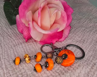 May include: A keychain with orange and yellow beads and a large orange circular bead. The keychain is attached to a silver clasp and ring. A pink and peach rose is in the background.