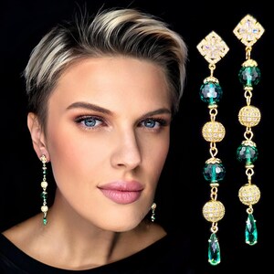 Puede incluir: Pendientes largos dorados con diseño colgante. Cada pendiente presenta una parte superior cuadrada con cristales transparentes, seguida de esferas doradas, cuentas verdes y colgantes verdes en forma de lágrima. Fondo negro.