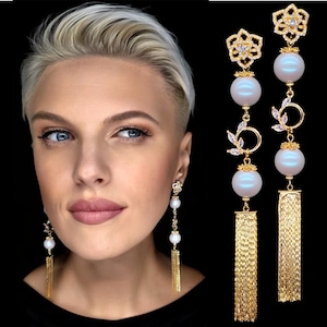 Puede incluir: Pendientes dorados con grandes cuentas de perlas y flecos dorados colgantes. Los pendientes presentan detalles florales y de hojas con cristales transparentes. El diseño está sobre un fondo negro.