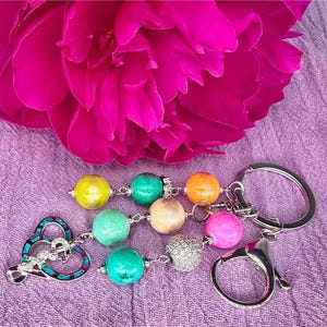 Puede incluir: Un llavero con cuentas de colores y un dije en forma de corazón. Las cuentas son en tonos amarillo, verde, naranja, rosa y crema. Una gran flor rosa vibrante en el fondo.