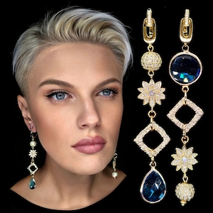 Puede incluir: Pendientes largos dorados con múltiples formas geométricas y florales, adornados con cristales transparentes y azules. Los pendientes presentan un cristal azul en forma de lágrima en la parte inferior y un cristal azul redondo en la parte superior. Sobre un fondo negro.
