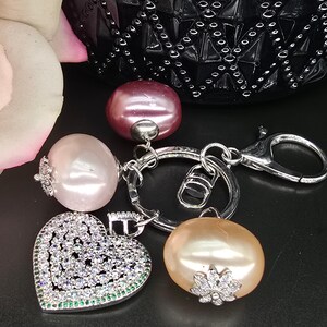 Op de afbeelding: Een zilveren sleutelhanger met een hartvormige hanger, een roze parel en twee kleinere roze parels. De hartvormige hanger is bedekt met heldere strass steentjes.