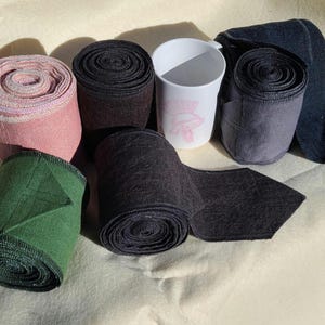 May include: An assortment of rolled fabric bandages in various colors, including pink, green, black, and gray. A white mug with a pink floral design is also present. The items are arranged on a light-colored surface.