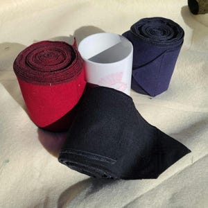 May include: An assortment of rolled fabric bandages in red, navy blue, and black, alongside a white ceramic mug with a pink floral design. The bandages are tightly wound, showcasing their texture and color.