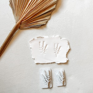 May include: A white rectangular clay slab featuring two identical embossed botanical sprig patterns, accompanied by two small rectangular white stamps with matching raised floral designs and a dried fan-shaped palm leaf.