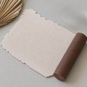 May include: A rectangular sheet of off-white textured paper with deckled edges, partially rolled up at one end. The rolled portion is a dark brown color with a visible grid-like texture, contrasting with the lighter, flatter paper section.