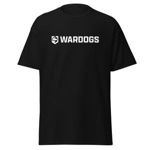 May include: A black short-sleeved t-shirt featuring a white graphic on the chest. The graphic consists of a shield icon containing a stylised wolf head silhouette, positioned to the left of the bold, capitalised white text that reads WARDOGS.