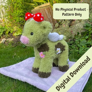 May include: Green crocheted plush toy cow with a red and white polka dot bow, brown hooves, and white wings. The cow is sitting on a light purple blanket with the text "Digital Download" in green on a yellow background.