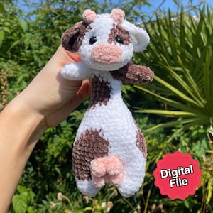 Puede incluir: Un peluche de vaca de ganchillo hecho a mano en blanco, marrón y rosa. La vaca tiene manchas, orejas y hocico marrones. El peluche tiene un aspecto suave y texturizado. La imagen incluye una pegatina roja que dice "Digital File".