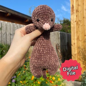 Könnte beinhalten: Ein braunes, plüschiges, handgemachtes Wesen mit rundem Kopf, kleinen Armen und einem bauchigen Körper. Das Wesen hat schwarze Augen und eine rosa Nase. Das Bild enthält einen roten Aufkleber mit der Aufschrift "Digital File".