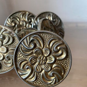 Könnte beinhalten: Ein Satz von fünf goldfarbenen Metall-Schubladengriffen mit floralem Design. Die Griffe sind rund und haben ein erhabenes, verziertes Muster.