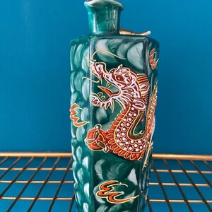 Könnte beinhalten: Eine grüne Keramikflasche mit einem detaillierten Drachendesign in Rot, Weiß und Gold. Die Flasche hat eine strukturierte Oberfläche und einen kleinen, goldfarbenen Verschluss. Die Flasche steht auf einem goldenen Drahtregal vor blauem Hintergrund.