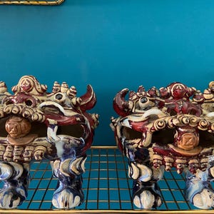 May include: Two ceramic guardian lion statues with intricate details. The statues are primarily cream, burgundy, and blue, with a textured finish. They are displayed on a gold wire shelf against a teal background.