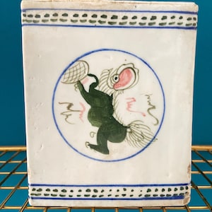 Puede incluir: Contenedor cuadrado de cerámica blanca con un diseño pintado en azul y verde. El contenedor presenta una imagen circular central de una criatura verde con boca roja y una red. Detalles de borde azules y verdes.