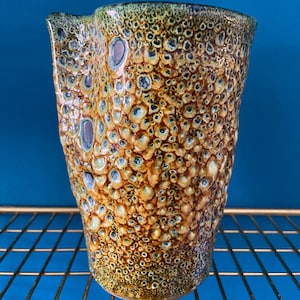 Puede incluir: Un jarrón de cerámica con una superficie texturizada y burbujeante en tonos marrones, amarillos y azules. El jarrón tiene una forma irregular y orgánica.