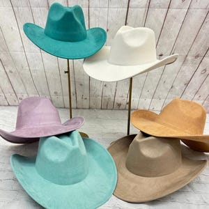 May include: A collection of six wide-brimmed hats in various colours including teal, white, lavender, light blue, tan, and beige. Each hat features a pinched crown and a curved brim, displayed on individual stands against a light wood background.