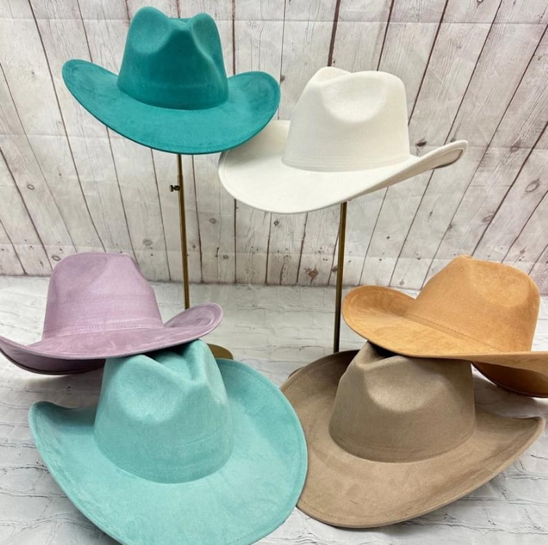 May include: A collection of six wide-brimmed hats in various colours including teal, white, lavender, light blue, tan, and beige. Each hat features a pinched crown and a curved brim, displayed on individual stands against a light wood background.