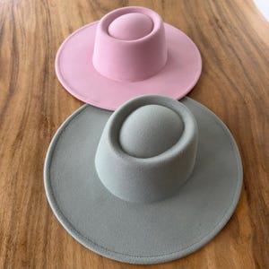 May include: Two felt hats, one in a light pink color and the other in a light gray color. Both hats have a wide brim and a tall, rounded crown.