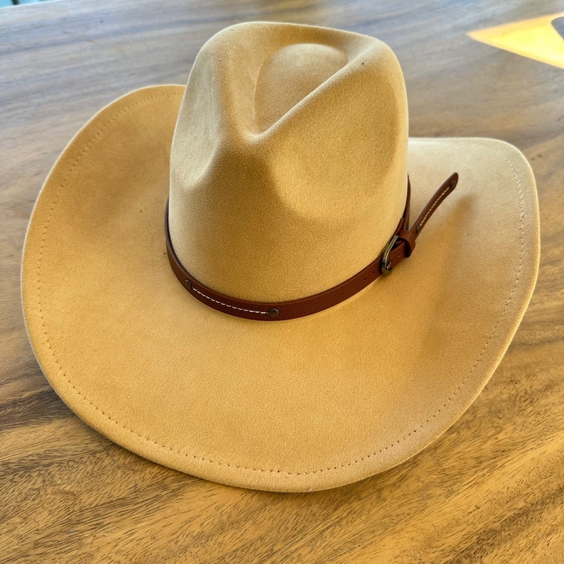 May include: A tan-colored cowboy hat with a wide brim and a brown leather band. The hat has a classic western design, perfect for sun protection. The leather band is secured with a metal buckle.