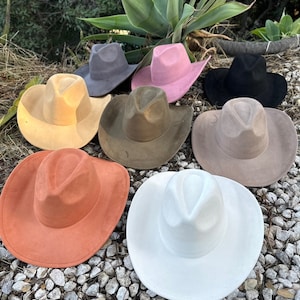 May include: A collection of eight felt cowboy hats in various colours including white, orange, tan, olive green, beige, grey, pink, and black. Each hat features a pinched crown and a wide, curved brim, arranged in a group on a rocky surface.