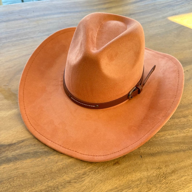 May include: A terracotta-colored cowboy hat with a brown leather band and buckle. The hat has a wide brim and a slightly curved crown. The leather band is stitched with white thread.