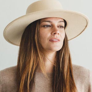 May include: A woman wearing a wide-brimmed, cream-colored felt hat with a tall crown. She is wearing a light brown sweater and has long brown hair.