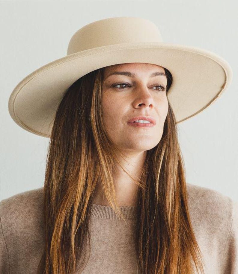 May include: A woman wearing a wide-brimmed, cream-colored felt hat with a tall crown. She is wearing a light brown sweater and has long brown hair.
