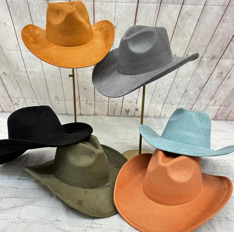 May include: A collection of six cowboy hats in various colours including orange, grey, black, olive green, and light blue. Each hat features a wide brim, a pinched crown, and a smooth, matte texture. The hats are displayed on individual stands.