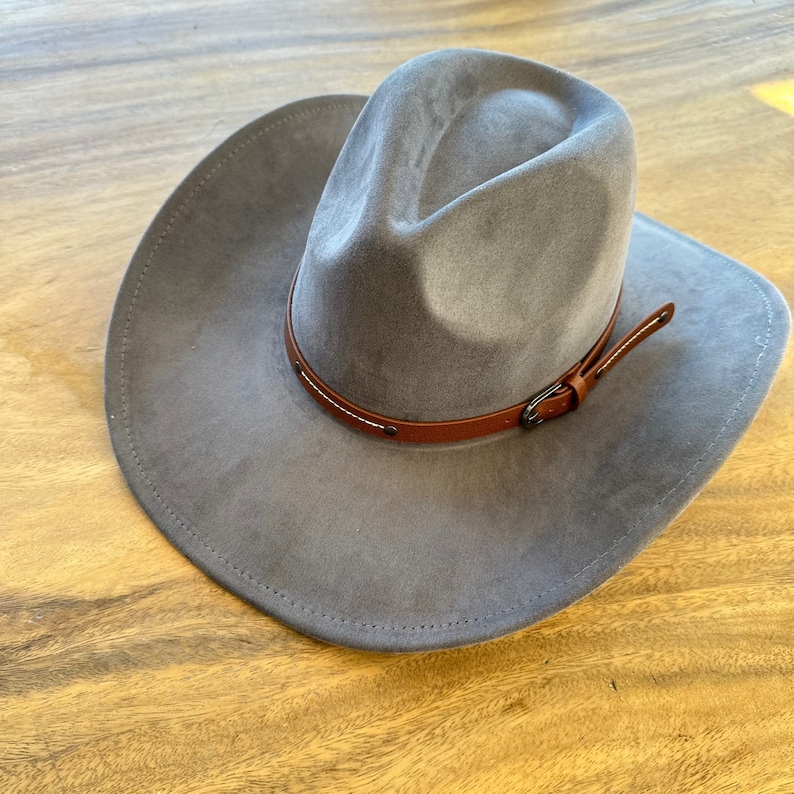 May include: A gray cowboy hat with a brown leather band and buckle. The hat has a wide brim and a rounded crown. The leather band is stitched with white thread. The hat is made of a soft, felt-like material.
