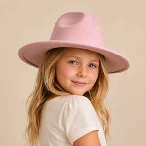 May include: A light pink fedora hat with a structured crown and a wide, flat brim. The hat features a smooth, felt-like texture and a matching tonal band around the base of the crown, providing a clean and uniform appearance in a soft pastel hue.