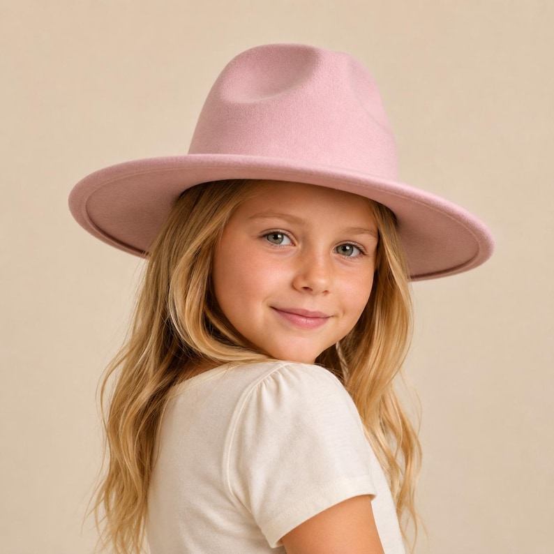 May include: A light pink fedora hat with a structured crown and a wide, flat brim. The hat features a smooth, felt-like texture and a matching tonal band around the base of the crown, providing a clean and uniform appearance in a soft pastel hue.